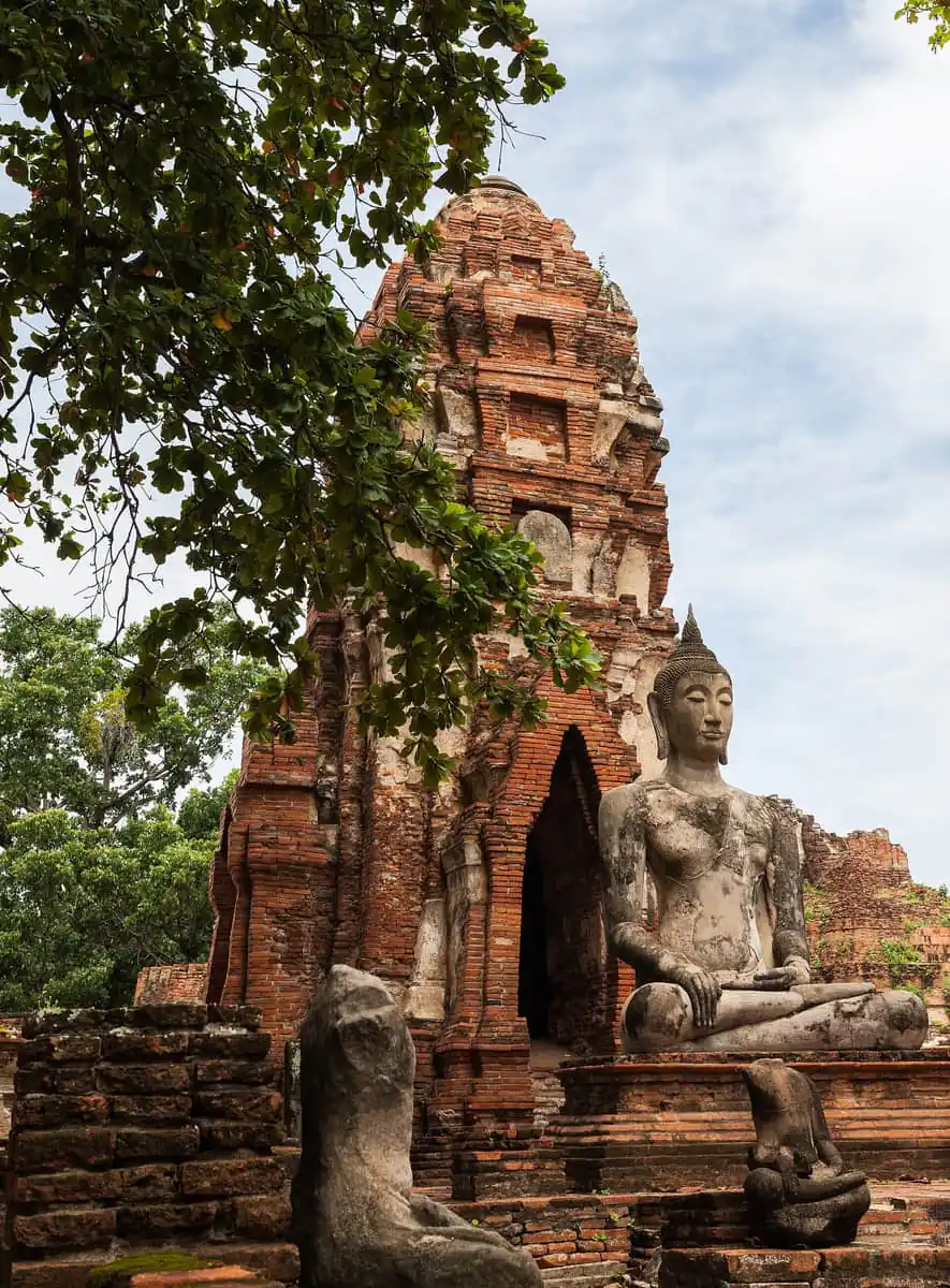 A seated Buddha statue in front of a brick prang at Wat Mahathat in Ayutthaya