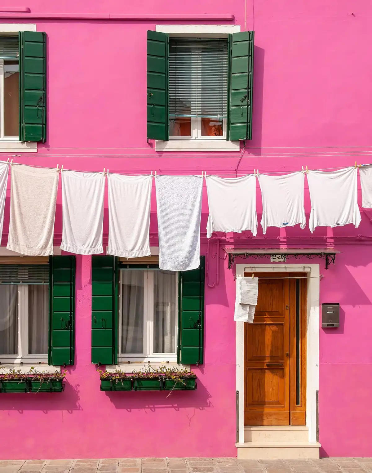 White laundry drying across a pink house front with green shutters on Burano