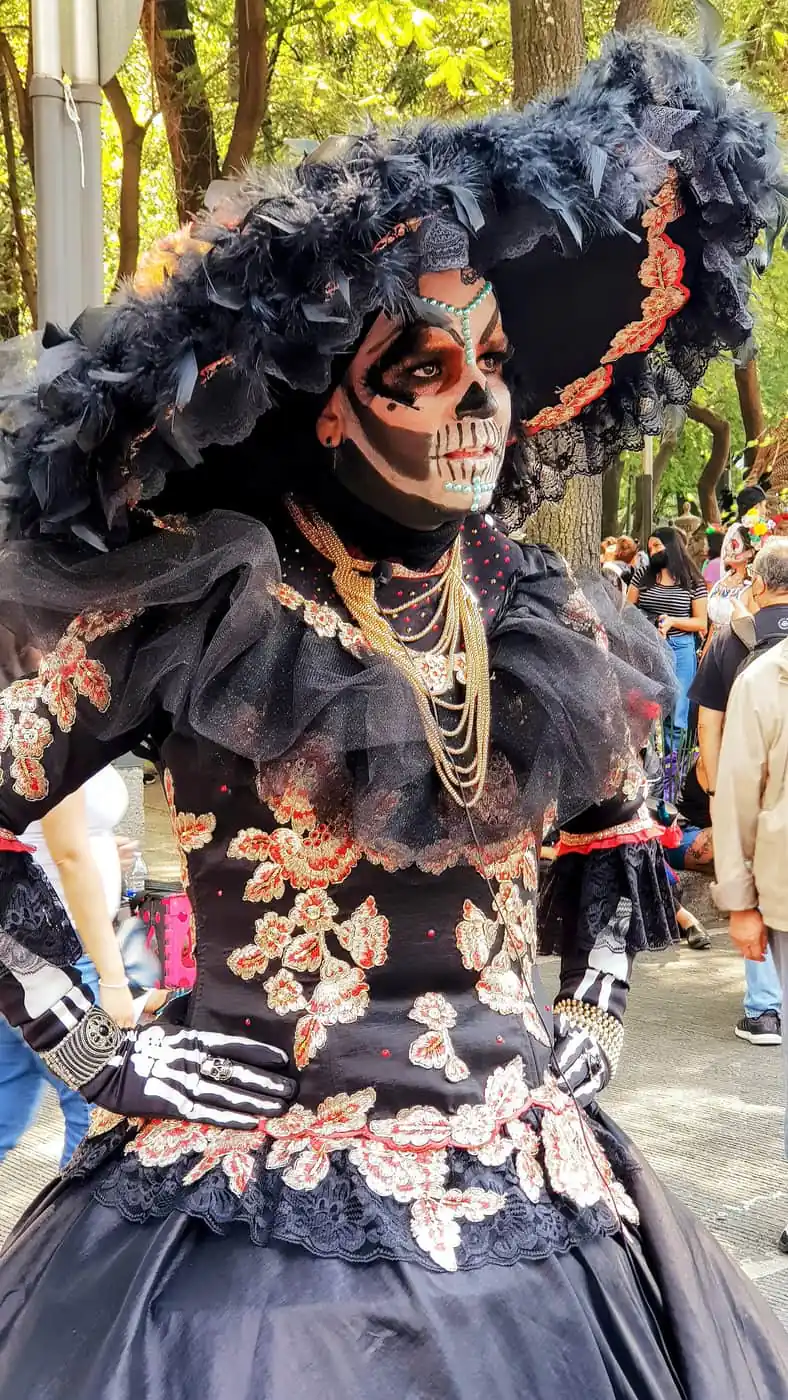 A Catrina costume with skull makeup, a feathered hat and an embroidered black dress at the Day of the Dead parade
