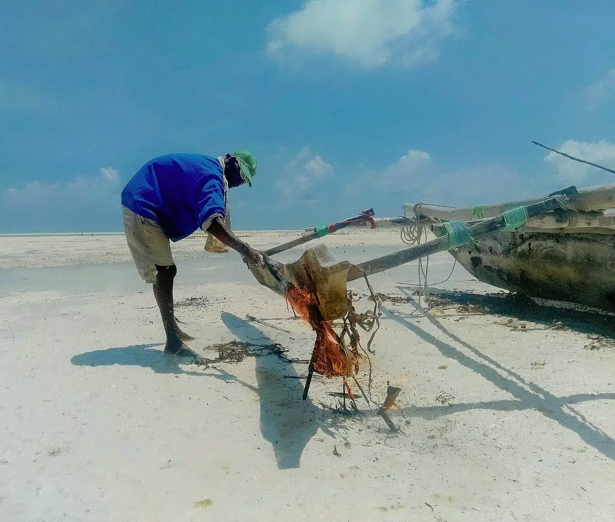 A fisherman tending a smoking dhow hull on white sand under blue sky