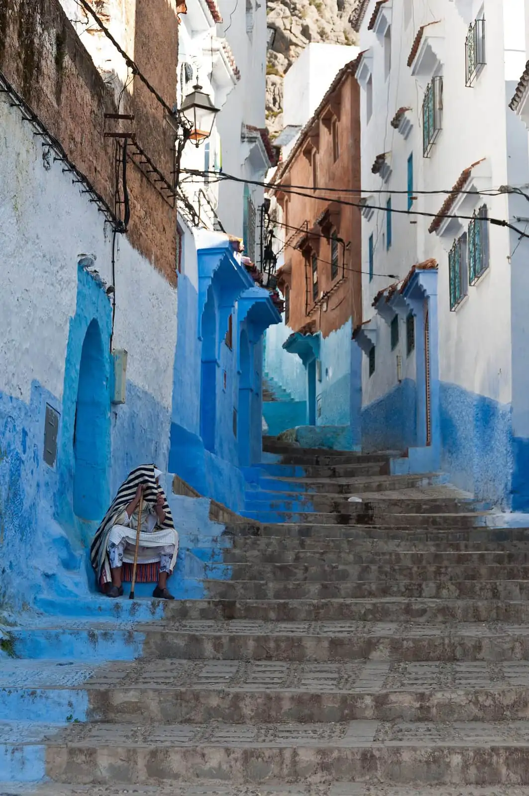 A stepped blue and white street in Chefchaouen with a seated figure in a striped cloak