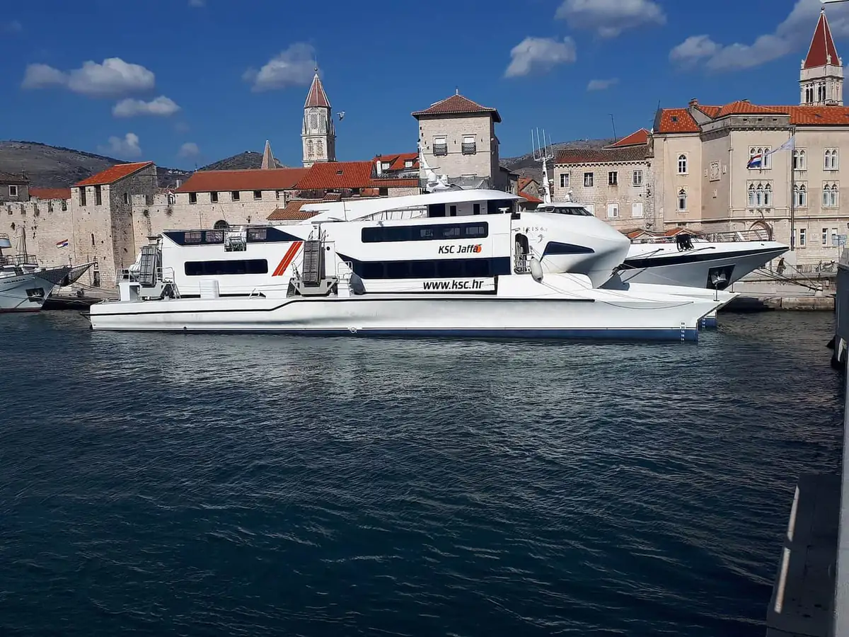 A white Krilo Shipping catamaran tied up at a stone quay in front of a walled old town