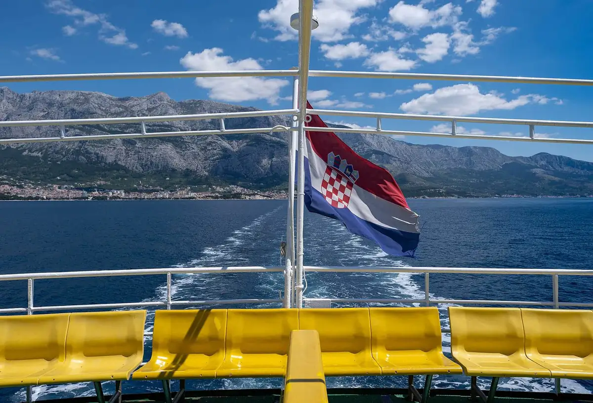 A Croatian flag flying from the stern rail of a ferry, with yellow deck seats and the Biokovo mountains behind Makarska