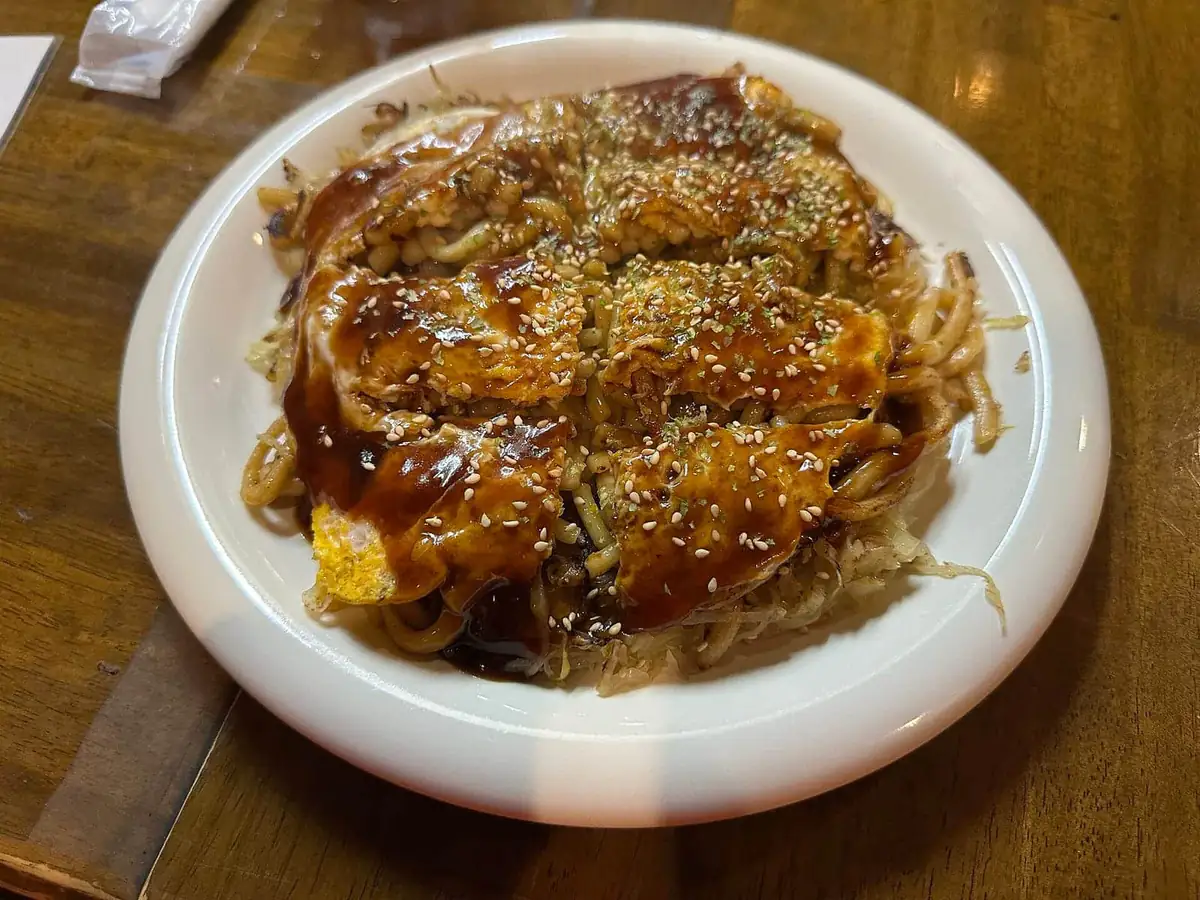A finished Hiroshima-style okonomiyaki of layered noodles, cabbage and sauce on a white plate