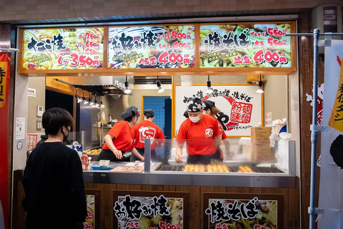 Staff cooking behind a takoyaki stand with illuminated menu boards in Shinsekai, Osaka