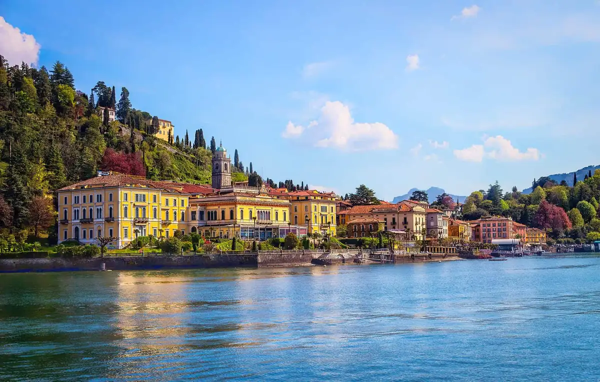 Bellagio waterfront on Lake Como seen from the water, with yellow villas, a church tower and mountains behind