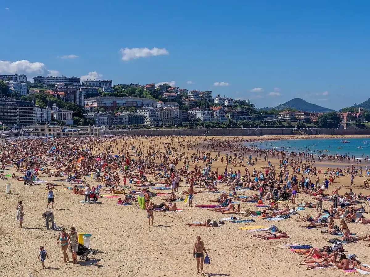La Concha beach in San Sebastián full of swimmers on a sunny summer day, with the bayfront promenade and green hills behind