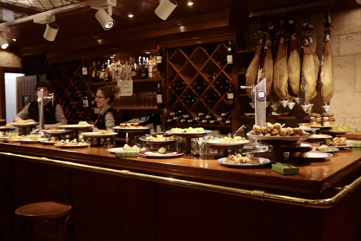 A pintxos bar counter in San Sebastián with plates of small bites along the bar and cured hams hanging behind