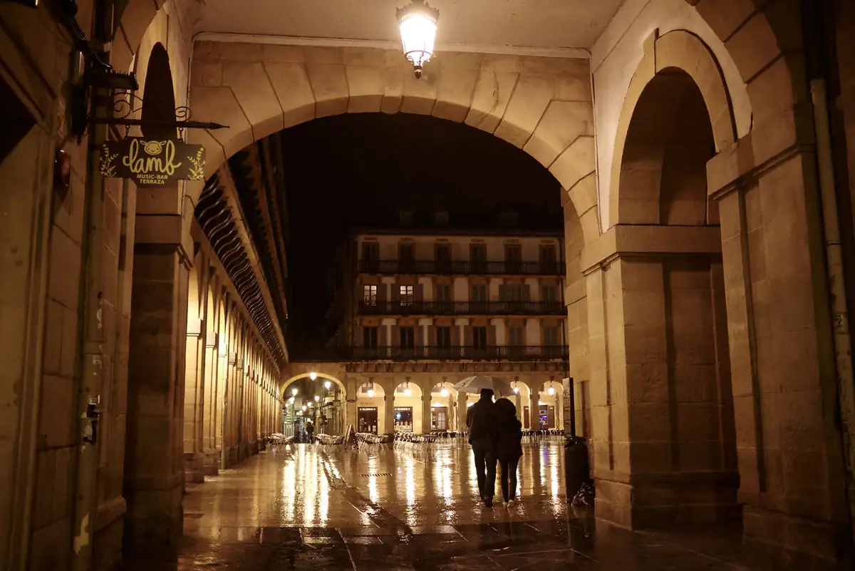 Plaza de la Constitución in San Sebastián at night in the rain, seen through the arcade with its balconied facades lit