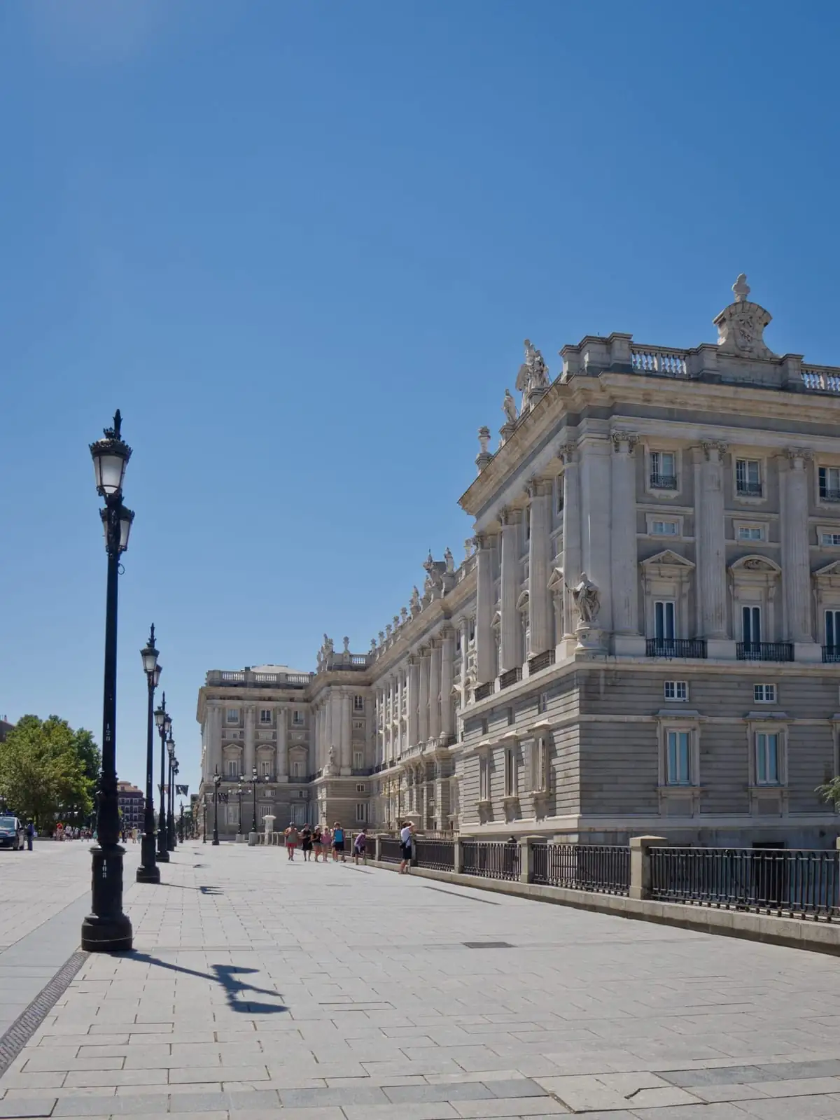 The stone facade of the Royal Palace of Madrid stretching along a wide paved plaza with street lamps