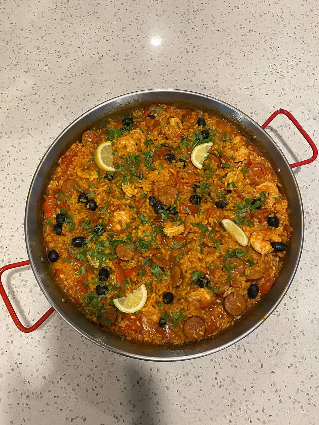 A pan of mixed seafood-style rice with prawns, lemon wedges and olives