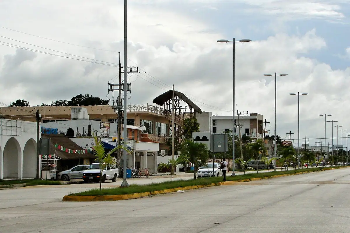 A cloudy day on a wide road through Tulum town, with low shops, palm trees and street lamps