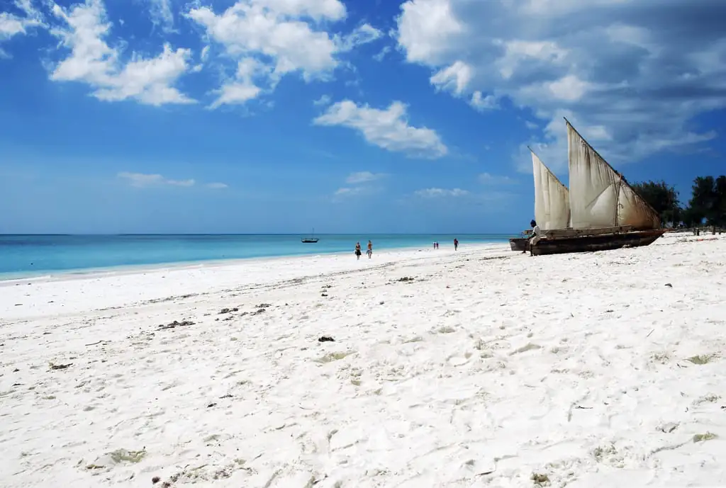 A dhow sailing boat pulled up on white sand with walkers on the beach